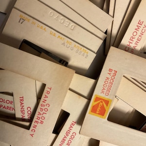 May include: A collection of vintage square cardboard photographic slide mounts in off-white. Each mount features red printed text reading Color Transparency, Kodak branding, and various stamped dates like DEC 70 and AUG 66A2, Made in U.S.A.