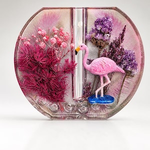 May include: A circular resin vase featuring a central glass tube, embedded with pink and purple dried flowers, and a small pink flamingo figurine standing on a blue base. The clear resin contains subtle glitter and spherical air bubbles.