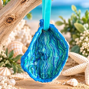 Könnte beinhalten: Ein dekoratives Austernschalen-Ornament mit handbemaltem Innenraum, der wellenförmige vertikale Streifen in den Farben Dunkelblau, Türkis und Meergrün aufweist. Die Schale ist an einer helltürkisfarbenen Satinschleife zum Aufhängen befestigt.