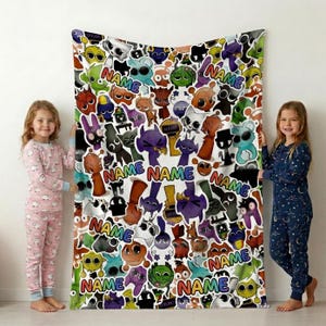 May include: A white fleece blanket featuring a colorful, repeating pattern of various cartoon creatures in shades of purple, orange, green, and blue. The design includes multiple instances of the word NAME in a vibrant, multi-colored rainbow font.