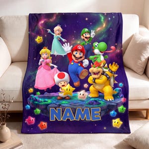 May include: A plush throw blanket featuring a vibrant space background with Mario, Luigi, Princess Peach, Rosalina, Yoshi, Bowser, and Toad. The characters are positioned on a floating island with colourful stars and the word NAME in blue text.