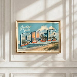 Puede incluir: Una impresión de arte mural horizontal enmarcada con el texto Greetings From DUCK en letras grandes. El diseño muestra una escena costera con una playa de arena, olas del mar, hierba de duna, un muelle de madera y un faro al atardecer.