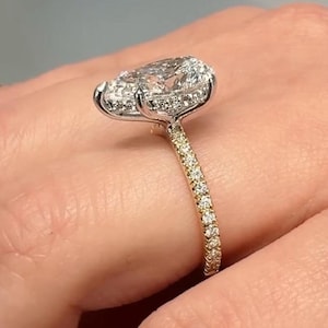 May include: A pear-shaped diamond ring featuring a large central stone set in a silver-toned metal basket. The band is crafted from a yellow-toned metal and is encrusted with a row of small, round, clear stones along the visible side of the shank.