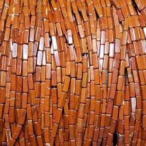 May include: Close-up of strands of reddish-brown rectangular beads. The beads have a polished, glossy finish and are strung together, creating a textured surface. The beads are uniform in shape and size, with slight variations in color.