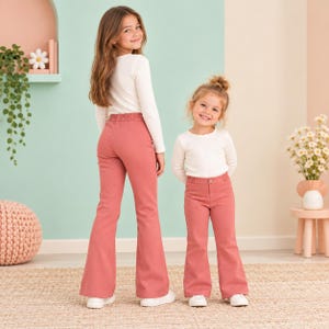 May include: Two individuals wearing long-sleeved white shirts and solid dusty rose flare pants. The taller individual is positioned on the left, facing away, while the shorter individual stands on the right, facing forward, both wearing white sneakers.