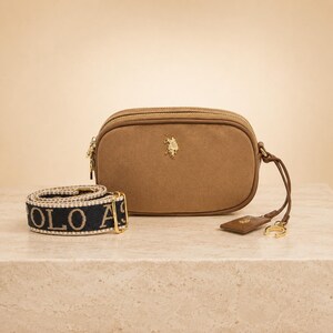 May include: A tan suede crossbody bag featuring a gold-toned polo player logo on the front. The bag includes a detachable navy blue and tan woven strap with the text POLO ASSN and a gold-toned zipper pull with a matching logo charm attached.