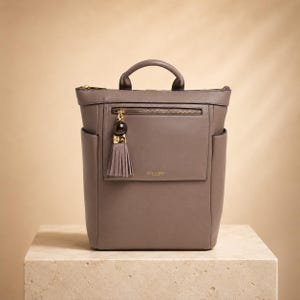 May include: A taupe leather backpack featuring a top handle, a zippered front pocket with a gold-toned tassel charm, and side slip pockets. The front pocket displays the gold embossed text Luella Grey London, and the bag has a structured silhouette.