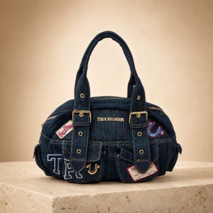 Könnte beinhalten: Eine dunkelblaue Jeans-Handtasche mit zwei Tragegriffen, goldenen Schnallen und Ösen. Die Tasche zeigt den Schriftzug True Religion, ein Hufeisen-Logo und verschiedene gestickte Aufnäher. Sie verfügt über zwei Vordertaschen und einen Reißverschluss.