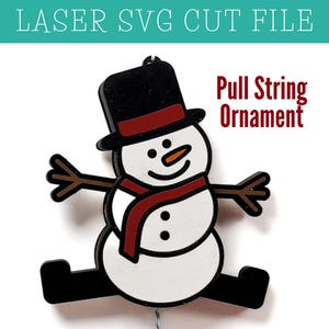 May include: A black and white snowman with a red scarf and black top hat. The snowman has a carrot nose and is holding out its arms. The text "Pull String Ornament" is written above the snowman.