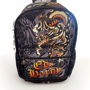May include: A black backpack featuring a large, detailed dragon graphic in shades of orange, yellow, and white. The front pocket displays the Ed Hardy logo in a gradient orange and yellow font. A small silver skull charm hangs from the zipper pull.