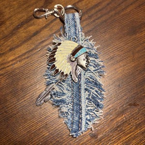 May include: A keychain featuring a frayed blue denim fabric strip shaped like a feather, attached to a silver-tone metal lobster clasp. A metal charm depicting a profile of a person wearing a feathered headdress is attached to the denim.
