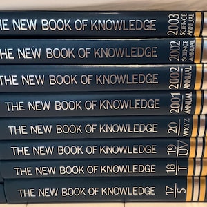 May include: A stack of dark blue hardcover books titled The New Book of Knowledge with gold lettering and Grolier logos on the spines. The volumes include various annuals and alphabetical sets ranging from 17/S to 2003 Science Annual.