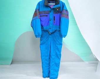 May include: A bright blue one-piece snowsuit featuring a hood, a front zipper closure, and a waist belt. The upper chest and sleeves are accented with purple panels and a multi-colored geometric pattern. The garment includes vertical zippered pockets.