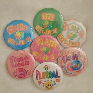 Könnte beinhalten: Sieben runde Ansteckbuttons mit farbenfrohen, handgezeichneten Texten und Illustrationen. Die Buttons zeigen Schriftzüge wie OSDD SYSTEM, DID SYSTEM, PLURAL SYSTEM, P-DID SYSTEM, FRONT STUCK, BLENDY und PLURRAL sowie verschiedene Smileys.