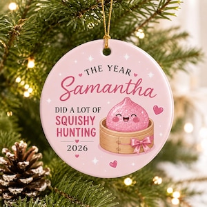 May include: A round, light pink ceramic ornament featuring a gold hanging string. The text reads THE YEAR Samantha DID A LOT OF SQUISHY HUNTING 2026. It displays a pink, smiling, squishy character inside a bamboo steamer basket with a pink bow.
