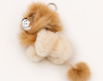 May include: A plush lion keychain featuring a fluffy tan mane, a cream-colored body, and a tail ending in a tan pom-pom. The lion has a white face with black embroidered details for the nose and mouth, and a silver metal ring attached to the top.