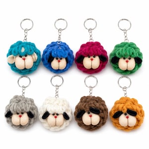 May include: Eight sheep keychains featuring round, textured yarn bodies in teal, blue, magenta, green, gray, white, brown, and orange. Each keychain has a beige fabric face, black ears, a small red nose, and a silver metal ring with a chain.