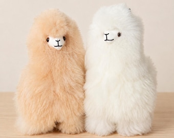 May include: Two plush alpaca toys with long, fluffy fur. One alpaca is light tan and the other is white. Both have small, dark brown eyes and embroidered black noses and mouths. The toys are standing upright, showcasing their soft, shaggy texture.