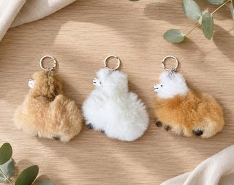 May include: Three fluffy llama keychains with metal rings. One is light brown, one is white, and one has a white head with a brown body. Each features a small, simple face with black eyes and a nose, crafted from soft, textured material.