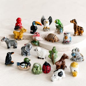 May include: A collection of small, hand-painted ceramic animal figurines including a toucan, eagle, parrot, giraffe, donkey, elephant, sheep, crocodile, ladybug, and various other creatures in vibrant colors, arranged in a group on a light surface.