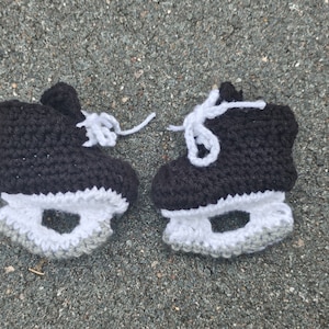 May include: A pair of black and white crocheted baby booties shaped like ice skates. The booties have white laces and a gray crocheted detail around the bottom.
