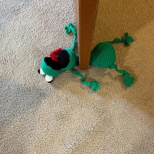 Op de afbeelding: Een groen gehaakt kikker speelgoed met zwarte en rode accenten. De kikker ligt op zijn rug met zijn poten in de lucht.