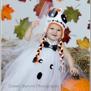 May include: A baby dressed as a ghost for Halloween, wearing a white tulle tutu and a crocheted hat with a Jack Skellington design. The baby is sitting next to a large orange pumpkin.