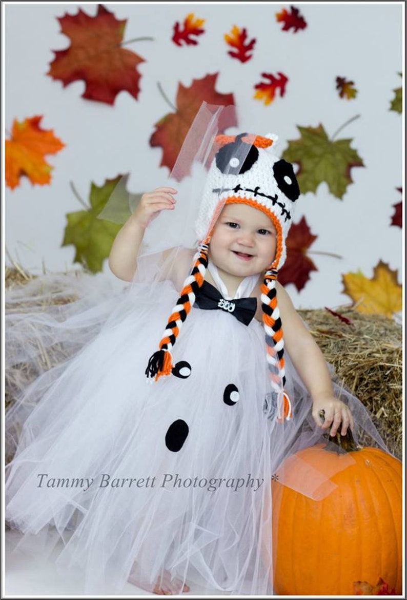 May include: A baby dressed as a ghost for Halloween, wearing a white tulle tutu and a crocheted hat with a Jack Skellington design. The baby is sitting next to a large orange pumpkin.