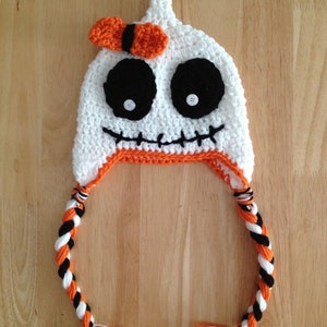 May include: White crocheted hat with a black and orange braided tail, featuring a black and white skull design with button eyes and a black stitched mouth. The hat has an orange and white bow on top.