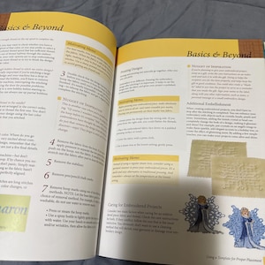 Puede incluir: Libro abierto titulado "Basics & Beyond" con páginas que muestran texto e ilustraciones sobre técnicas de bordado. Las páginas son amarillas y blancas, con un fondo azul. El libro está abierto a una sección sobre el cuidado de proyectos de bordado.