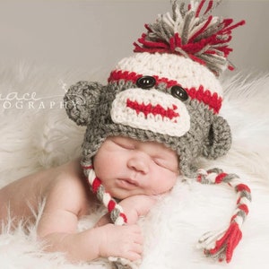 May include: A baby wearing a gray and white crocheted monkey hat with red accents and a long tail. The hat has a pom-pom on top and the baby is sleeping on a white fluffy surface.