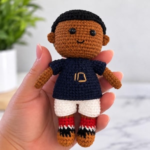 Può includere: Una piccola bambola all'uncinetto con carnagione bruna, capelli neri e occhi a perla neri. La bambola indossa una maglietta blu navy con il numero 10 ricamato in oro, pantaloncini bianchi e calzini rossi, bianchi e neri con scarpe marroni.