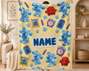 Pode incluir: Uma manta de lã amarela com um padrão repetitivo de Blue de Blue's Clues, poltronas vermelhas, lápis de cera roxos, cadernos azuis, despertadores e molduras. O centro exibe a palavra NAME em letras maiúsculas azuis.