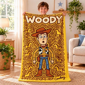 Könnte beinhalten: Eine gelbe, rechteckige Fleecedecke mit einer großen Cartoon-Illustration der Figur Woody aus Toy Story. Das Design zeigt den Schriftzug WOODY oben und Cowboy unten, umgeben von braunen Kritzellinien in Form von kleinen Pferden.