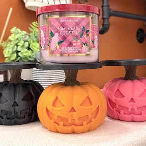 May include: Three jack-o'-lantern candle pedestals in black, orange, and pink, each featuring a carved face and a brown stem base. A Bath & Body Works Mrs. Claus' Cocktail candle in a clear jar with a pink plaid label sits on the centre pedestal.