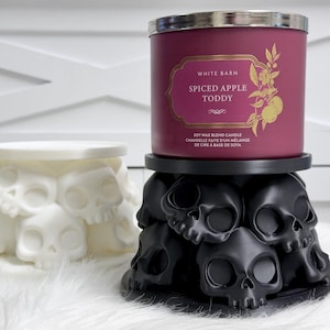 Peut inclure: Une bougie cylindrique avec une étiquette rouge foncé portant l'inscription White Barn Spiced Apple Toddy, posée sur un socle noir en forme de crânes. À côté se trouve un socle identique blanc. Les deux socles présentent des crânes moulés.