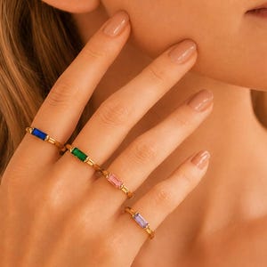 Puede incluir: Cuatro anillos de tono dorado, cada uno con una piedra preciosa rectangular de corte baguette. Las piedras están dispuestas en fila y muestran colores distintos: azul profundo, verde vibrante, rosa suave y morado claro, sobre bandas finas.