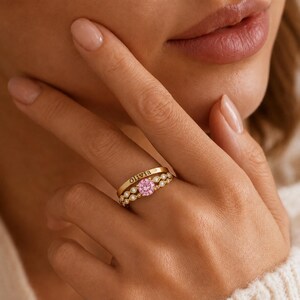 Puede incluir: Un conjunto de anillos de oro que incluye una banda con el nombre Olivia grabado en negro y otra banda con una piedra preciosa rosa redonda, rodeada por un diseño metálico festoneado con pequeñas piedras transparentes brillantes.