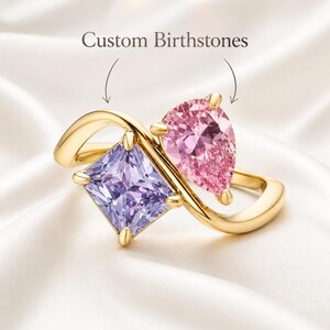 Puede incluir: Un anillo de oro con dos piedras: una piedra morada de corte cuadrado y una piedra rosa de corte en forma de pera. La banda tiene un diseño cruzado. Sobre el anillo, el texto Custom Birthstones aparece con dos flechas hacia las gemas.