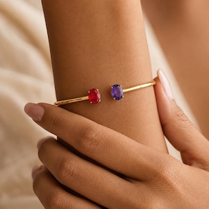 Puede incluir: Una pulsera abierta de tono dorado con dos piedras ovaladas, una de color rojo vibrante y otra de color morado intenso, situadas en cada extremo de la banda. La banda de metal es lisa y pulida, creando un diseño sencillo y limpio en la muñeca.