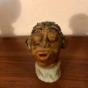 May include: A hand-sculpted ceramic figurine of a head with a glossy brown glaze, textured hair, closed eyes, and red-painted lips. The head rests on a cylindrical base featuring a light-colored finish with subtle, repeating vertical line patterns.