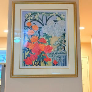 May include: A framed print featuring a vibrant garden scene with orange, red, and pink roses climbing a black wrought iron gate. A yellow vase sits on a pedestal beside white flowers, all set against a light blue background in a gold frame.