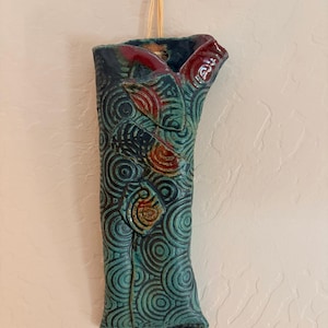 May include: A wall-hanging ceramic vase with a teal and dark blue glaze featuring a repeating circular pattern. The vase has three raised, leaf-shaped decorative elements with red and orange accents and is suspended by a light-colored raffia string.