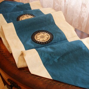 May include: A teal table runner with a cream border and gold embroidered circles. The runner has a tassel on one end.