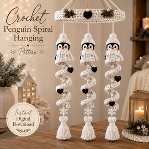 May include: A crochet hanging mobile featuring three white and black penguin figures wearing hats and scarves, suspended from a circular frame. The design includes spiral elements, small black hearts, and white snowflake accents, finished with tassels.