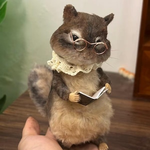 Könnte beinhalten: Eine kleine, flauschige Eichhörnchen-Figur in Braun und Creme, die aufrecht steht. Das Eichhörnchen trägt eine dünne, runde Brille aus kupferfarbenem Draht und einen zarten, cremefarbenen Spitzenkragen. Es hält ein winziges, offenes Buch.