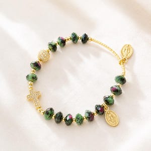May include: A beaded bracelet featuring faceted green and magenta zoisite beads, small gold-tone spacer beads, a gold-tone pave cross charm, a gold-tone pave sphere bead, and two oval gold-tone religious medals with embossed figures.