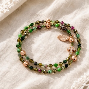 May include: A multi-strand wrap bracelet featuring faceted green and olive-toned crystal beads, small metallic rose gold spheres, and three charms: a metallic rose gold rose, a textured leaf, and a small cross encrusted with clear crystals.