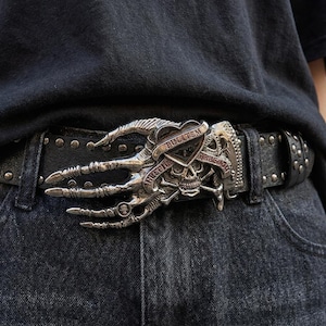 May include: A black leather belt featuring a large silver-toned metal buckle shaped like a skeletal hand clutching a skull and a heart. The heart is engraved with the text PULLTEM and the skull is positioned above crossed bones on the belt buckle.