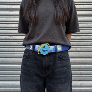 Peut inclure: Une ceinture bleue ornée d'un motif répétitif d'icônes informatiques et d'une grande boucle circulaire en forme de logo Internet Explorer. La ceinture est portée avec un t-shirt noir uni et un jean noir, mettant en valeur un design graphique.